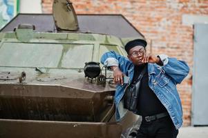 afroamerikanischer mann in jeansjacke, baskenmütze und brille, mit zigarre, die gegen ein gepanzertes btr-militärfahrzeug posiert. foto