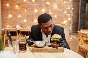 respektabler junger Afroamerikaner in schwarzem Anzug und Handschuhen zum Essen, der im Restaurant mit leckerem Doppelburger und Sodagetränk sitzt. foto