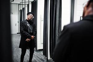 Stilvoller lässiger afroamerikanischer Mann mit schwarzer Baskenmütze und Mantel mit Gürteltasche im Umkleidekabinen-Bekleidungsgeschäft, Blick auf den Spiegel. foto