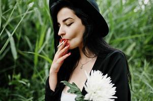 sinnliches rauchermädchen ganz in schwarz, roten lippen und hut. goth dramatische frau hält weiße chrysanthemenblume und raucht auf gemeinem schilf. foto