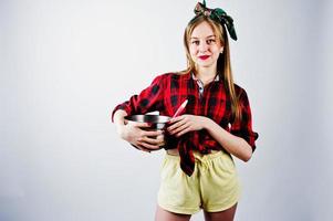 junge lustige Hausfrau in kariertem Hemd und gelben Shorts Pin-up-Stil mit Topf und Küchenlöffel isoliert auf weißem Hintergrund. foto