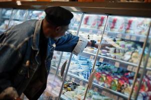 Stilvoller lässiger afroamerikanischer Mann in Jeansjacke und schwarzer Baskenmütze, der einen Korb hält, in der Nähe eines gastronomischen Kühlschranks steht und im Supermarkt einkauft. foto