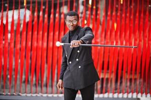 Stilvoller afroamerikanischer Gentleman in eleganter schwarzer Jacke, der einen Retro-Spazierstock als Rohrkolben oder einen Kippstock mit goldenem Diamantkugelgriff hält. reicher modischer afro-mann gegen rote wand. foto