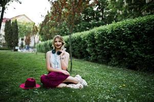 Modisches und schönes blondes Modellmädchen in stylischem rotem Samt-Veloursrock, weißer Bluse und Hut, sitzend auf grünem Gras im Park mit Telefon und Kopfhörern. foto