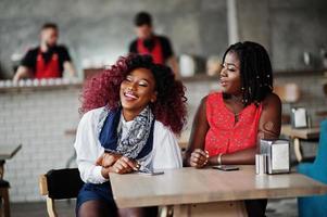 Attraktive afroamerikanische zwei Freundinnen, die mit Mobiltelefonen am Tisch im Café sitzen.
