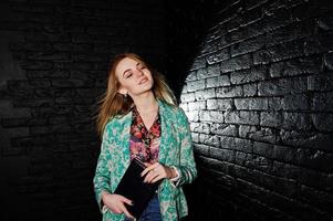 stylisches blondes Mädchen in Jacke und Jeans mit Notizbuch-Tagebuch in der Hand vor schwarzer Ziegelwand im Studio. foto