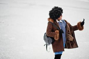 lockiges haar afroamerikanerin trägt schaffellmantel und handschuhe mit rucksack, der am wintertag gestellt wird. foto