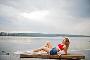 sexy lockiges Model-Mädchen in rotem Oberteil, Jeans-Denim-Shorts, Brille und Turnschuhen posiert auf einer Bank am See. foto