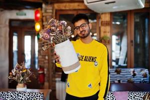 asiatischer mann in brillen und gelbem sweatshirt posiert im innencafé und hält die vase mit getrockneten blumen. foto