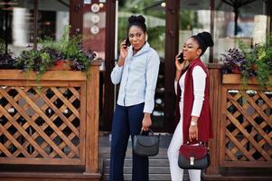 Zwei stylische afroamerikanische Freundinnen mit Handtaschen posierten im Freien der Straßenstadt und sprachen am Handy. foto