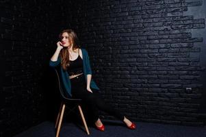 hübsche brünette mädchen tragen eine schwarze und grüne jacke in roten high heels, sitzen und posieren auf einem stuhl im studio gegen eine dunkle backsteinmauer. Studio-Model-Porträt. foto