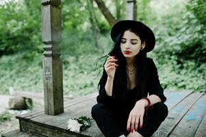 sinnliches rauchermädchen ganz in schwarz, roten lippen und hut. goth dramatische frau, die dünne zigarette raucht. foto