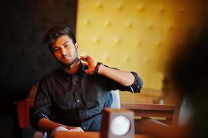Selbstbewusster junger Inder im schwarzen Hemd, der im Café sitzt. foto