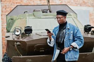 afroamerikanischer mann in jeansjacke, baskenmütze und brille, der am telefon gegen ein gepanzertes btr-militärfahrzeug spricht. foto