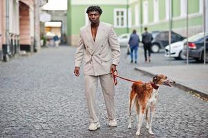 Stilvoller Afro-Mann im beigen Old-School-Anzug mit russischem Barsoi-Hund. Modischer junger afrikanischer Mann in Freizeitjacke auf nacktem Oberkörper. foto