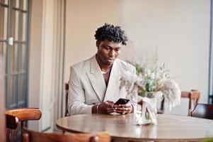 Stilvoller Afro-Mann im beigefarbenen Anzug der alten Schule, der mit Handy im Café sitzt. Modischer junger afrikanischer Mann in Freizeitjacke auf nacktem Oberkörper. foto