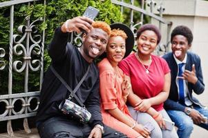 vier afrikanische freunde haben spaß im freien. zwei schwarze mädchen mit jungs mit handys machen selfie. foto