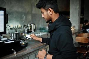 Lässiger und stilvoller junger asiatischer Mann mit Kopfhörern im Café an der Bar, der per Kreditkarte am POS-Terminal bezahlt. foto