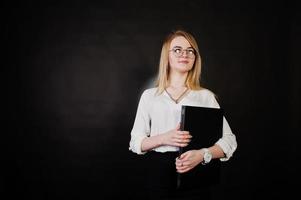 Studioporträt einer blonden Geschäftsfrau mit Brille, weißer Bluse und schwarzem Rock, die einen Laptop vor dunklem Hintergrund hält. erfolgreiche frau und stilvolles mädchenkonzept. foto