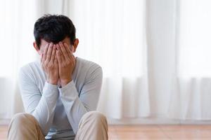 depressiver asiatischer junger mann, der gesicht mit händen bedeckt, allein zu hause weint, verärgerter sitzender mann, sich nach streit oder trennung unglücklich fühlt, verzweifelt und einsam und panik, psychologisches problemkonzept foto