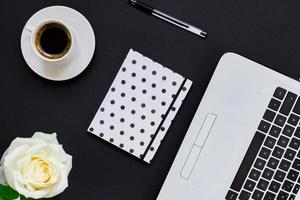 flache Lage, Draufsicht Bürotisch Schreibtisch. Arbeitsplatz mit Laptop, weißer Rose, gepunktetem Tagebuch und Kaffeebecher auf schwarzem Hintergrund. foto