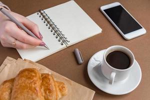 Frauenhand mit Stiftschreiben auf Notizbuch im Café. freiberufler, der im freien arbeitet. Kaffeepause mit Croissant foto