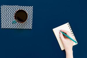 Blick von oben auf die Tasse schwarzen Kaffee und die Hand der Frau, die in einem Notizbuch auf blauem Hintergrund mit Kopierraum schreibt. foto