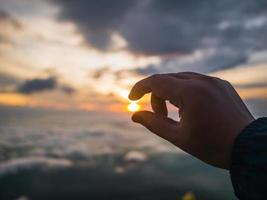 menschliche hand, die versucht, die sonne bei einem wunderschönen sonnenaufgang mit idyllischem wolkenhimmel am morgen auf dem berg aufzunehmen foto