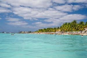 menschen schwimmen in der nähe von weißem sandstrand mit sonnenschirmen, bungalowbar und kokospalmen, türkisfarbenes karibisches meer, isla mujeres insel, karibisches meer, cancun, yucatan, mexiko foto