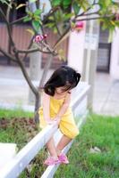 vertikales Foto eines asiatischen Mädchens im Alter von 3 Jahren, das einen weißen Zaun erklimmt, um auf die andere Seite zu springen. Das Kind trägt ein gelbes Kleid. das Konzept der Klettersicherheit.