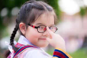 Schulmädchen, das die Uniform des jungen Mädchens trägt. Das Kind drückt mit dem Zeigefinger auf die Brille, die es trägt. süßes Lächeln. Zopf kleines Mädchen. trägt eine rot karierte Schuluniform. Ein asiatisches Mädchen ist 6 Jahre alt foto