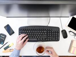 Draufsicht auf männliche Hände, die am Computer arbeiten und eine Teetasse auf dem Schreibtisch halten.