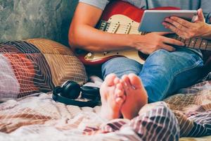 Nahaufnahme eines jungen Mannes, der ein Tablet benutzt und auf seinem Bett E-Gitarre spielt, Filme mit Kornfilter. foto