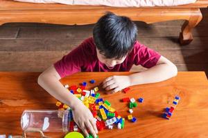 asiatischer junge wird kreativ, indem er bunte plastiksteine zu robotern und flugzeugen auf einem holztisch zusammenbaut, glücklich und lustig zu hause. foto