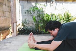 junger asiatischer mann, der yoga praktiziert, in sitzender vorwärtsbeugeübung sitzt, paschimottanasana-pose auf der grünen yogamatte, draußen im garten trainiert. gesunder Lebensstil und gesundes Wohnkonzept foto