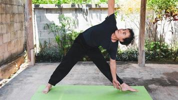 gesundes lebenskonzept des jungen asiatischen mannes, der yoga-asana-dreieckshaltung praktiziert, trainiert, posiert auf einer grünen yogamatte. Bewegung im Freien im Garten. gesunder Lebensstil. foto