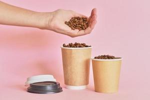 weibliche hand streut kaffeebohnen in kaffeepapierbecher auf rosa hintergrund foto