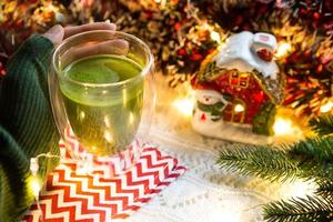 die hand der frau hält einen doppelwandigen glasbecher mit japanischem matcha-tee auf dem tisch mit weihnachtsdekor. Silvesterstimmung, Girlande und Lametta, Fichtenzweig, kuschelige, gestrickte Decke, gestreifte Serviette foto