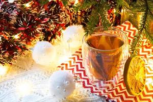 transparenter doppelwandiger glasbecher mit heißem tee und zimtstangen auf dem tisch mit weihnachtsdekor. Silvester-Atmosphäre, getrocknete Orangenscheibe, Girlande und Lametta, Fichtenzweig, gemütlich foto