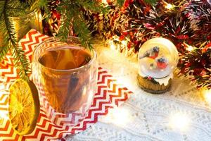 transparenter doppelwandiger glasbecher mit heißem tee und zimtstangen auf tisch mit weihnachtsdekor. Silvesterstimmung, getrocknete Orangenscheibe, Girlande und Lametta, Schneekugel mit Dompfaffen foto