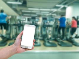 Hand, die ein mobiles Smartphone mit unscharfem Fitness-Studio-Gerätehintergrund, Vintage-Filter hält foto