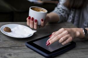 Frauenhand, die auf Tablette stöbert und Kaffee hält