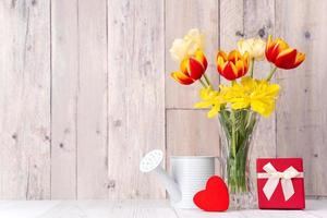 Tulpenblumenarrangement in Glasvase mit Herzgruß, Gießkannendekor auf Holztischhintergrundwand, Nahaufnahme, Muttertagsdesignkonzept. foto