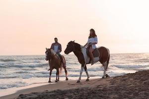 Ein liebevolles Paar in Sommerkleidung, das bei Sonnenuntergang an einem Sandstrand reitet. Meer und Sonnenuntergang im Hintergrund. selektiver Fokus foto