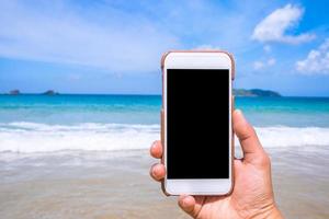 tourist, der telefon am strand mit dem meer verwendet, hand, die weißes mobiles smartphone smartphone, reisearbeitskonzept, unscharfer hintergrund, nahaufnahme hält.
