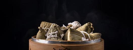 Reisknödel, Zongzi - Drachenbootfest, Haufen chinesischer traditioneller gekochter Speisen im Dampfgarer auf Holztisch auf schwarzem Hintergrund, Nahaufnahme, Kopierraum foto