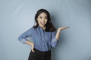 Aufgeregte asiatische Frau mit blauem Hemd, die auf den Kopierbereich neben ihr zeigt, isoliert durch blauen Hintergrund foto