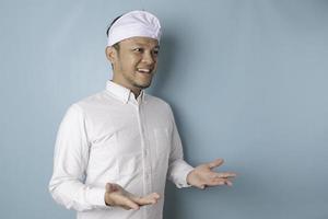 Junger männlicher Arzt, der eine Idee mit Udeng oder traditionellem Stirnband und weißem Hemd vorstellt, während er lächelnd auf isoliertem blauem Hintergrund aussieht foto