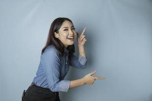 Aufgeregte asiatische Frau mit blauem Hemd, die auf den Kopierbereich neben ihr zeigt, isoliert durch blauen Hintergrund foto