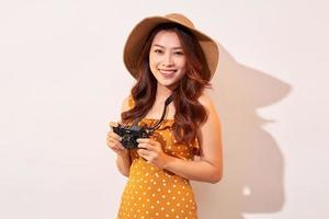 Porträt einer fröhlich lächelnden jungen Frau, die inspirierend fotografiert und ein Sommerkleid trägt. Mädchen mit Retro-Kamera. Model posiert auf beigem Hintergrund mit Hut foto